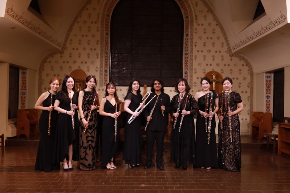 A flute choir in black, holding their flutes in an ornate hall
